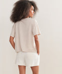 A woman with voluminous curly hair stands with her back to the camera, wearing the Vintage Pocket Tee in beige and white shorts, her hands tucked in her pockets against a plain light background.