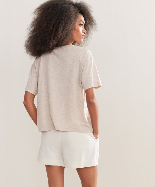 A woman with voluminous curly hair stands with her back to the camera, wearing the Vintage Pocket Tee in beige and white shorts, her hands tucked in her pockets against a plain light background.
