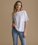 A woman modeling a white short-sleeve crewneck pocket t-shirt with light wash blue jeans, standing against a solid beige studio background.