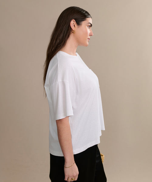 A woman with long brown hair stands in profile against a beige background, wearing the Vintage Pocket Tee in white, paired with black pants and gold jewelry.