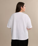 A woman with long brown hair, wearing the Vintage Pocket Tee and black pants, stands facing away from the camera against a plain beige background.