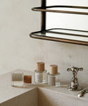 Three glass bottles with wooden caps, including Violet Santal Eau De Parfum with woody and floral notes, are displayed on a beige bathroom counter near a silver faucet, with a mirror and shelf above.