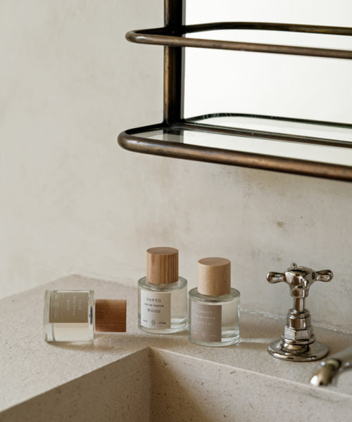 Three glass bottles with wooden caps, including Violet Santal Eau De Parfum with woody and floral notes, are displayed on a beige bathroom counter near a silver faucet, with a mirror and shelf above.