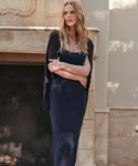 A woman with long wavy blonde hair stands before a stone fireplace, wearing the Rib Tank Dress in dark navy with a black sweater over her shoulders. She crosses her arms and gazes to the side, embodying effortless warm-weather style.