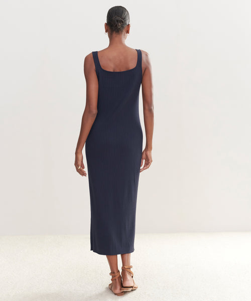 A woman faces away, wearing the Rib Tank Dress in dark navy with brown sandals. She stands in a minimal, light-filled space with a neutral background—showcasing this warm-weather staple.