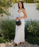 A woman stands outdoors on a gravel path, wearing a white Rove Ankle Dress with black polka dots, black sandals, and holding a small black handbag. Green bushes and a white fence are in the background.