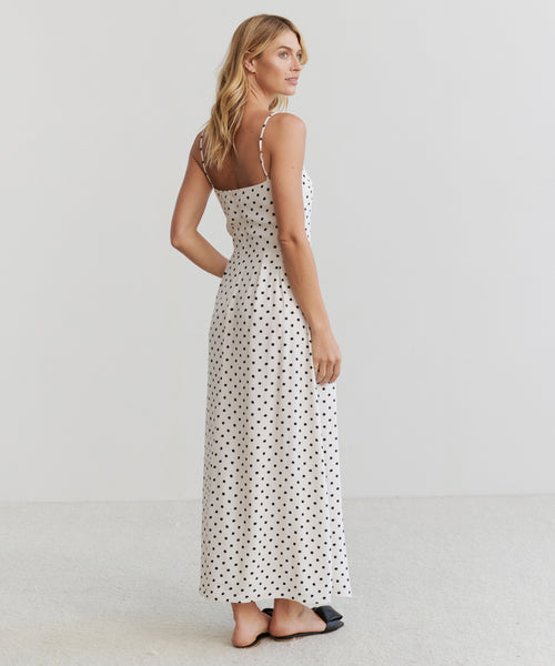A woman stands barefoot on a light carpet, wearing the Rove Ankle Dress—an ankle-length dress with black polka dots and thin shoulder straps. She faces away from the camera, looking over her shoulder against a plain light background.