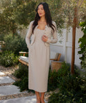 A woman stands outdoors on a stone path, wearing the Rove Dress in light beige. She pairs it with a matching cardigan, her long dark hair flowing, surrounded by greenery, a wooden bench, and a white fence in the background.