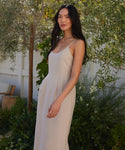 A woman with long dark hair stands outside by a white fence and green foliage, wearing the sleeveless light beige Rove Dress, made from fine Italian fabric, gazing calmly at the camera.