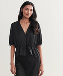 A woman with long, wavy dark hair wears the Sawyer Blouse—vintage-inspired, black Italian viscose with a V-neck, puff sleeves, and drawstring waist—paired with a black skirt against a plain light background.