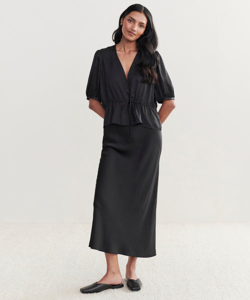 A woman poses against a plain light background, wearing the Sawyer Blouse in black with short puff sleeves and a waist tie, paired with a black ankle-length skirt and black flat shoes.