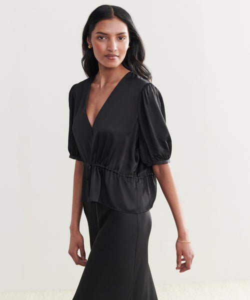 A woman with long dark hair wears a black Sawyer Blouse with short sleeves, V-neck, and elastic waist, paired with a matching skirt as she stands against a light background.