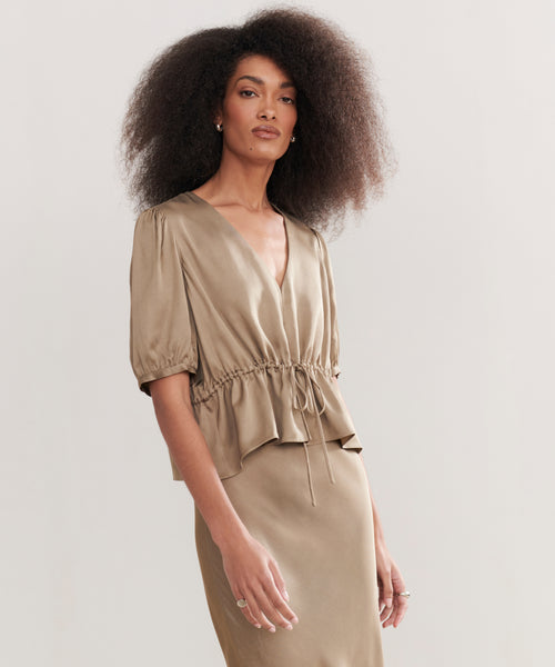 A woman with voluminous, curly hair wears the taupe Sawyer Blouse in Italian viscose—featuring short puff sleeves and a drawstring waist—with a matching skirt, standing against a plain light background.