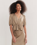 A woman with voluminous curly hair wears the Sawyer Blouse, a vintage-inspired taupe Italian viscose top with short sleeves, V-neckline, gathered waist, and matching skirt, standing against a plain light background.