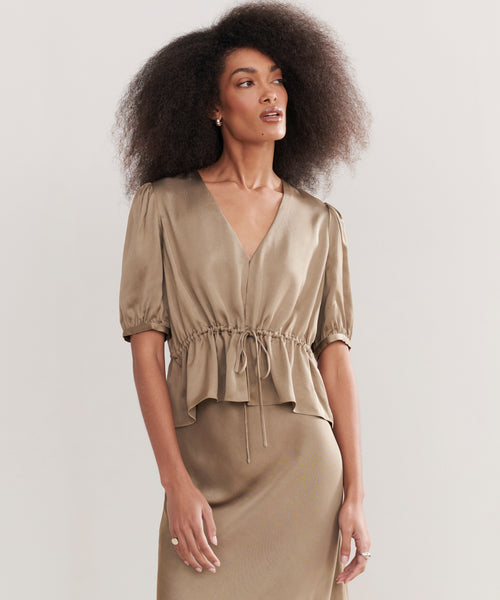A woman with voluminous curly hair wears the Sawyer Blouse, a vintage-inspired taupe Italian viscose top with short sleeves, V-neckline, gathered waist, and matching skirt, standing against a plain light background.