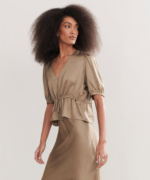 A woman with voluminous curly hair wears the Sawyer Blouse in light brown Italian viscose, with short sleeves, a V-neck, and a drawstring waist, paired with a matching skirt against a plain light background.