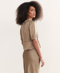 A woman with voluminous curly hair stands sideways, looking over her shoulder, wearing the vintage-inspired Sawyer Blouse in taupe Italian viscose against a plain light background.