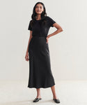 A woman with long dark hair and a confident posture stands against a plain light background, wearing the Sawyer Slip Skirt—a long black viscose skirt—paired with a short-sleeved black top and black flat shoes.