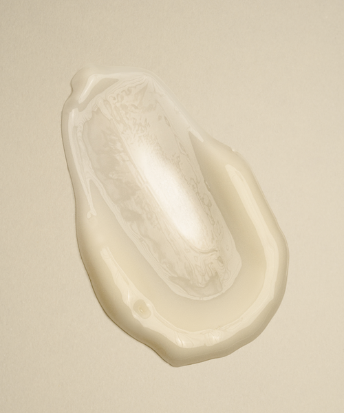 A close-up of a creamy, translucent, oval smear of Daily Vitamin C Serum on a pale beige surface, designed to brighten skin and target dark spots.