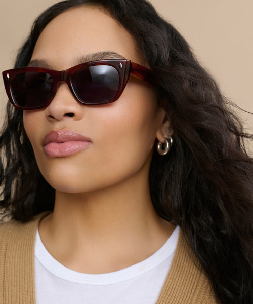 A woman with long wavy hair wears Webster Sunglasses, a white top, a tan cardigan, and silver hoop earrings, posing against a beige background.