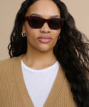 A woman with long wavy dark hair wears Webster Sunglasses, silver hoop earrings, a white shirt, and a tan knit cardigan against a beige background.
