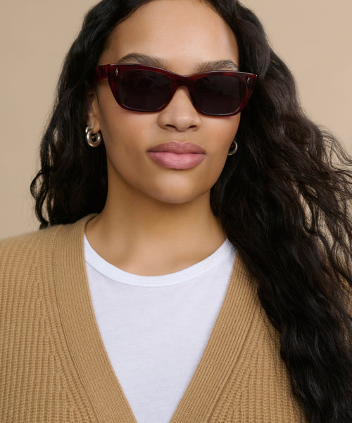 A woman with long wavy dark hair wears Webster Sunglasses, silver hoop earrings, a white shirt, and a tan knit cardigan against a beige background.