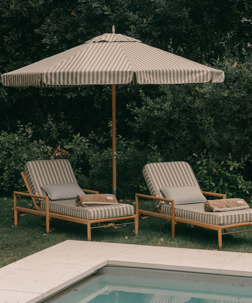 Two striped lounge chairs with cushions and a matching umbrella by a poolside.