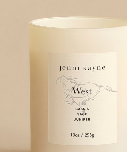 The West Glass Candle is a 10oz (295g) white natural wax candle in a frosted jar labeled jenni kayne West, featuring a minimalist horse illustration and California-inspired notes of cassis, sage, and juniper.