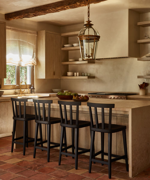 A warm, rustic kitchen with black oak cabinets, a large island with three Hampton Counter Stools, open shelves, a farmhouse sink, terracotta tile flooring, and a glass lantern-style pendant light.