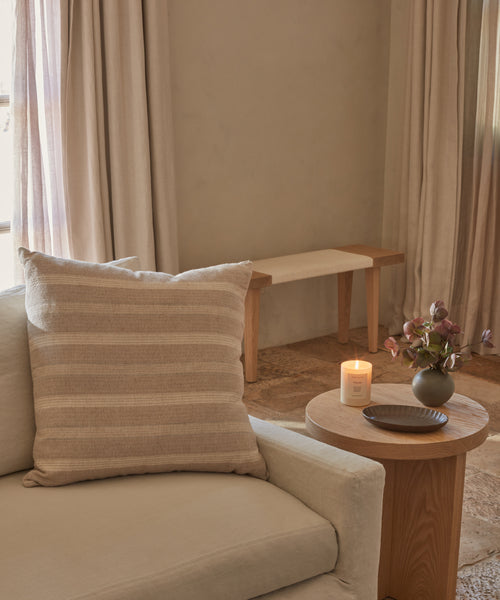 A cozy living room with a beige sofa, striped pillow, round wooden side table holding a lit candle, small dish, and vase with purple flowers. Light curtains and the Oak Day Bench are in the background.
