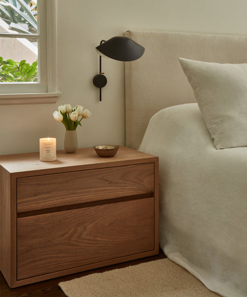 The Palms Nightstand holds a lit candle, a small vase of white tulips, and a bowl beside a bed with cream bedding. A black wall lamp is mounted above the handmade Palms Nightstand near a window with green leaves outside.