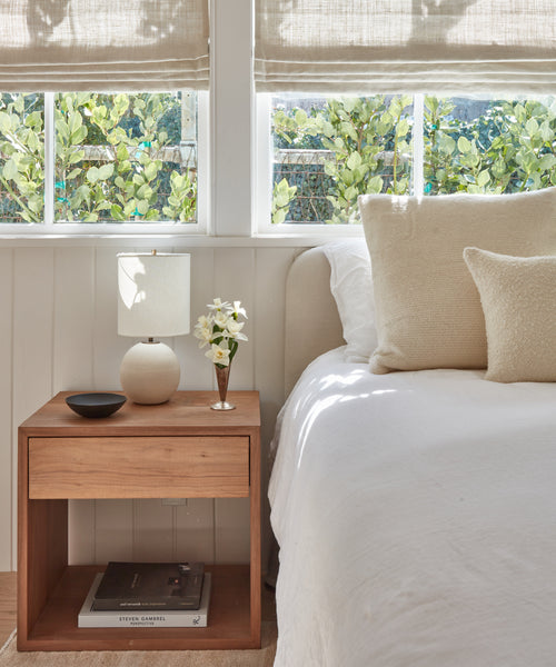 A cozy bedroom corner showcases the Berkeley Nightstand in solid walnut, topped with a lamp, a white flower vase, and a black dish next to a tidy bed. Sunlight filters through beige-shaded windows with leafy plants outside.