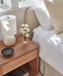 The Berkeley Nightstand displays a white lamp, white flowers in a vase, and a black dish beside a bed with white bedding, illuminated by sunlight from a nearby window.