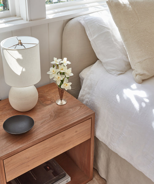 The Berkeley Nightstand displays a white lamp, white flowers in a vase, and a black dish beside a bed with white bedding, illuminated by sunlight from a nearby window.