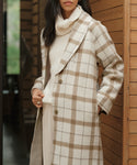 A woman stands indoors near a wooden wall, one hand in her pocket, wearing a cream turtleneck sweater and the Austin Wool Coat—a long oatmeal plaid coat with a shawl collar. Her face is partially out of view.