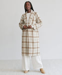 A person wearing the Austin Wool Coat in oatmeal plaid with a shawl collar over a white top and wide-legged white pants stands on a light background. They have long braided hair and tan shoes.
