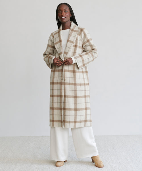A person wearing the Austin Wool Coat in oatmeal plaid with a shawl collar over a white top and wide-legged white pants stands on a light background. They have long braided hair and tan shoes.