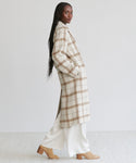 A woman in profile wears the Austin Wool Coat—a long, oatmeal plaid wool coat with a shawl collar—over white pants and tan heeled boots. She has long braided hair and poses against a plain light background.