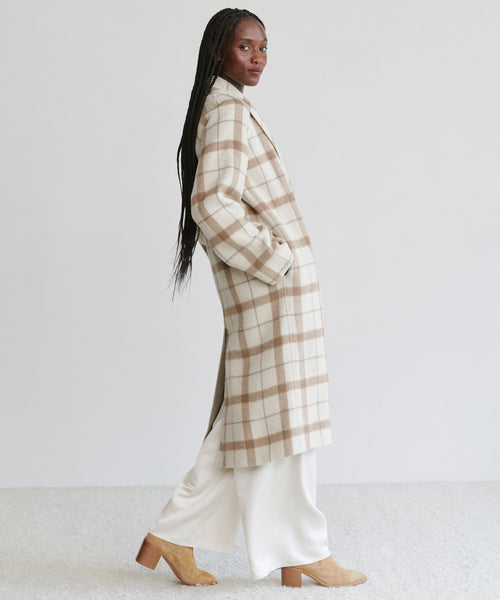 A woman in profile wears the Austin Wool Coat—a long, oatmeal plaid wool coat with a shawl collar—over white pants and tan heeled boots. She has long braided hair and poses against a plain light background.