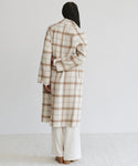 A person stands facing away from the camera, wearing the Austin Wool Coat—a long oatmeal plaid wool coat with a shawl collar—over cream-colored pants. Their long braided hair cascades down their back against a plain background.