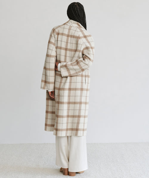 A person stands facing away from the camera, wearing the Austin Wool Coat—a long oatmeal plaid wool coat with a shawl collar—over cream-colored pants. Their long braided hair cascades down their back against a plain background.