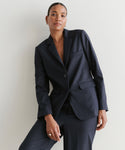 A woman with short hair and small hoop earrings wears the Brentwood Blazer in dark navy with matching trousers, standing confidently against a light background, showcasing the classic tailoring of her look.