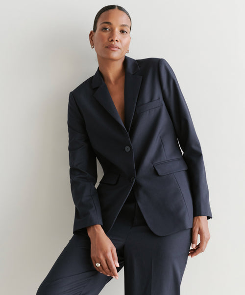 A woman with short hair and small hoop earrings wears the Brentwood Blazer in dark navy with matching trousers, standing confidently against a light background, showcasing the classic tailoring of her look.