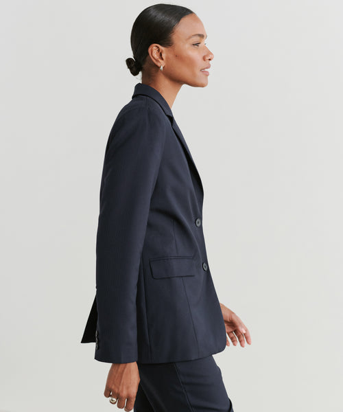 A woman in a dark navy Brentwood Blazer and matching pants stands in profile against a plain light background, her hair neatly tied back and rings on her fingers—showcasing the timeless style of the Brentwood Blazer.