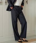 Wearing classic tailoring, this person styles the dark navy Brentwood Trouser with a white shirt, black belt, and black heels, standing indoors with one hand in their pocket against white paneled cabinets.