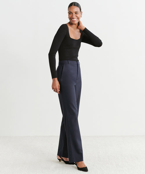 A woman stands confidently against a white background, wearing a fitted black long-sleeve top, black heels, and the Brentwood Trouser in dark navy, showcasing classic tailoring and effortless style.