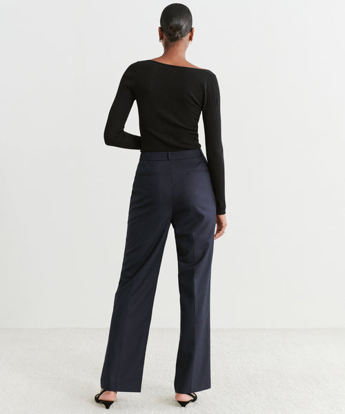 A woman stands with her back to the camera, hair in a sleek bun, wearing a fitted black long-sleeve top, Brentwood Trouser in dark navy, and black heels against a plain light background.