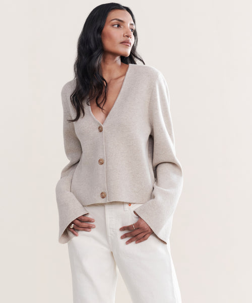 A woman with long dark hair wears the Chelsea Cardigan in light beige, featuring wide sleeves, paired with white pants. She stands against a plain light background, gazing to the side with one hand in her pocket.