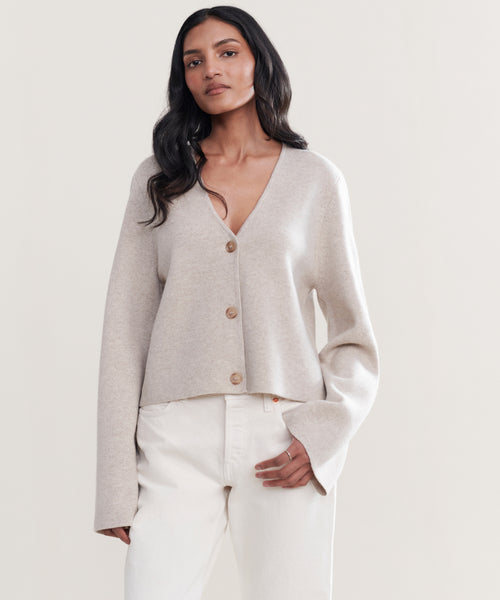 A woman with long dark hair models the Chelsea Cardigan, paired with high-waisted off-white pants, against a plain light background.