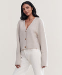 A woman with long dark hair wears the Chelsea Cardigan in light beige with brown buttons and off-white pants, standing against a plain, light background.